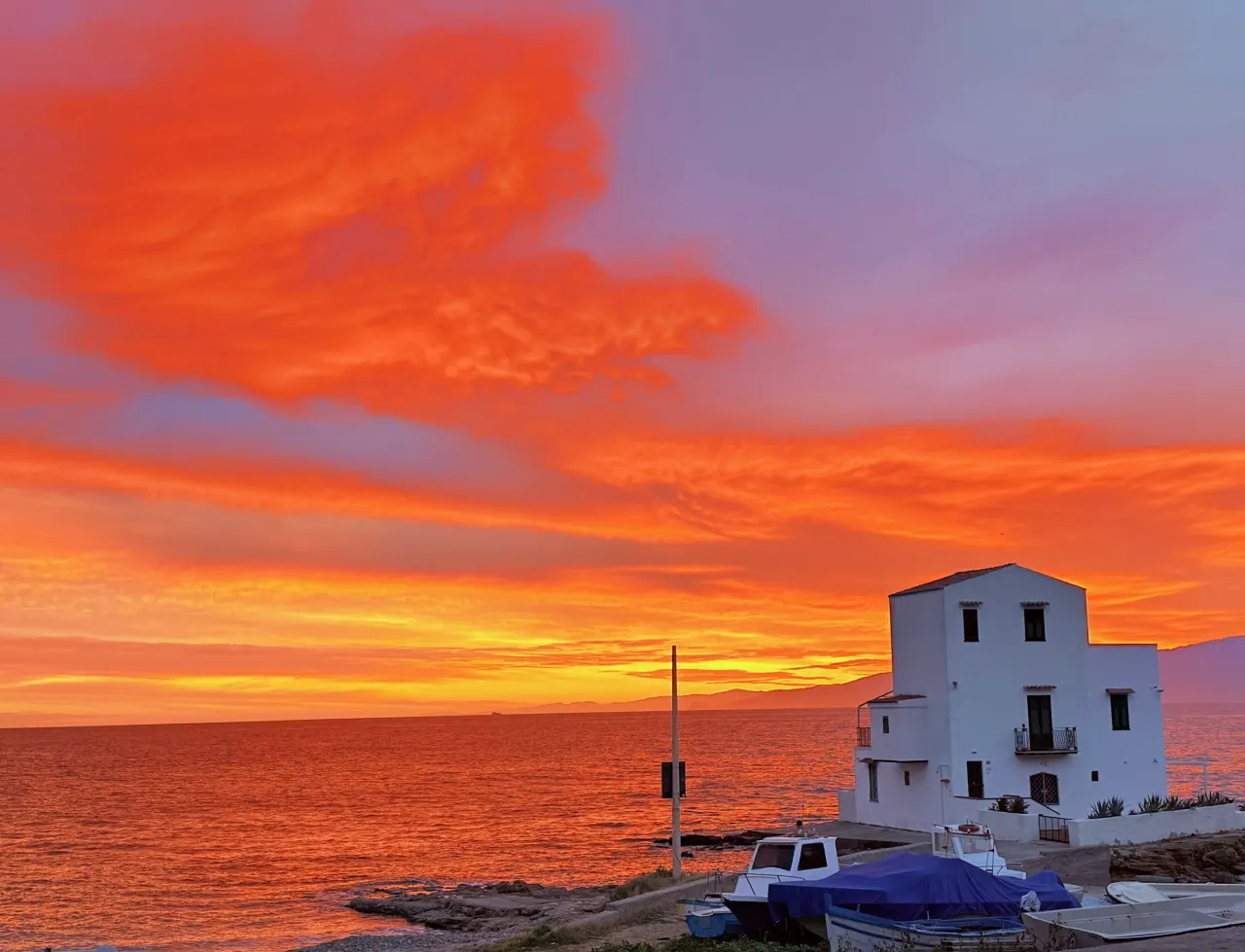 Sonnenuntergang über dem Meer, am Ufer ein weißes Haus und Fischerboote
